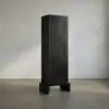 Tall black wooden cabinet with three square-panel doors on a chunky base, standing in a minimalist room.