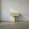 Minimalist light wood cabinet with a shallow drawer on a round pedestal against a pale wall.