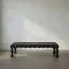 Low, black wooden bench with carved legs placed on a concrete floor against a light textured wall